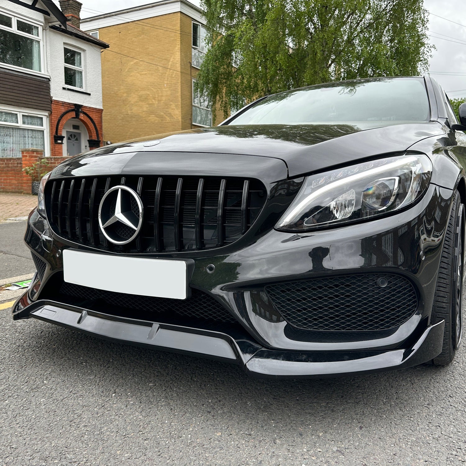 Car parked on a residential street, featuring a glossy black finish and a prominent logo on the front grille. Surrounded by brick and cream houses with visible driveway and greenery.