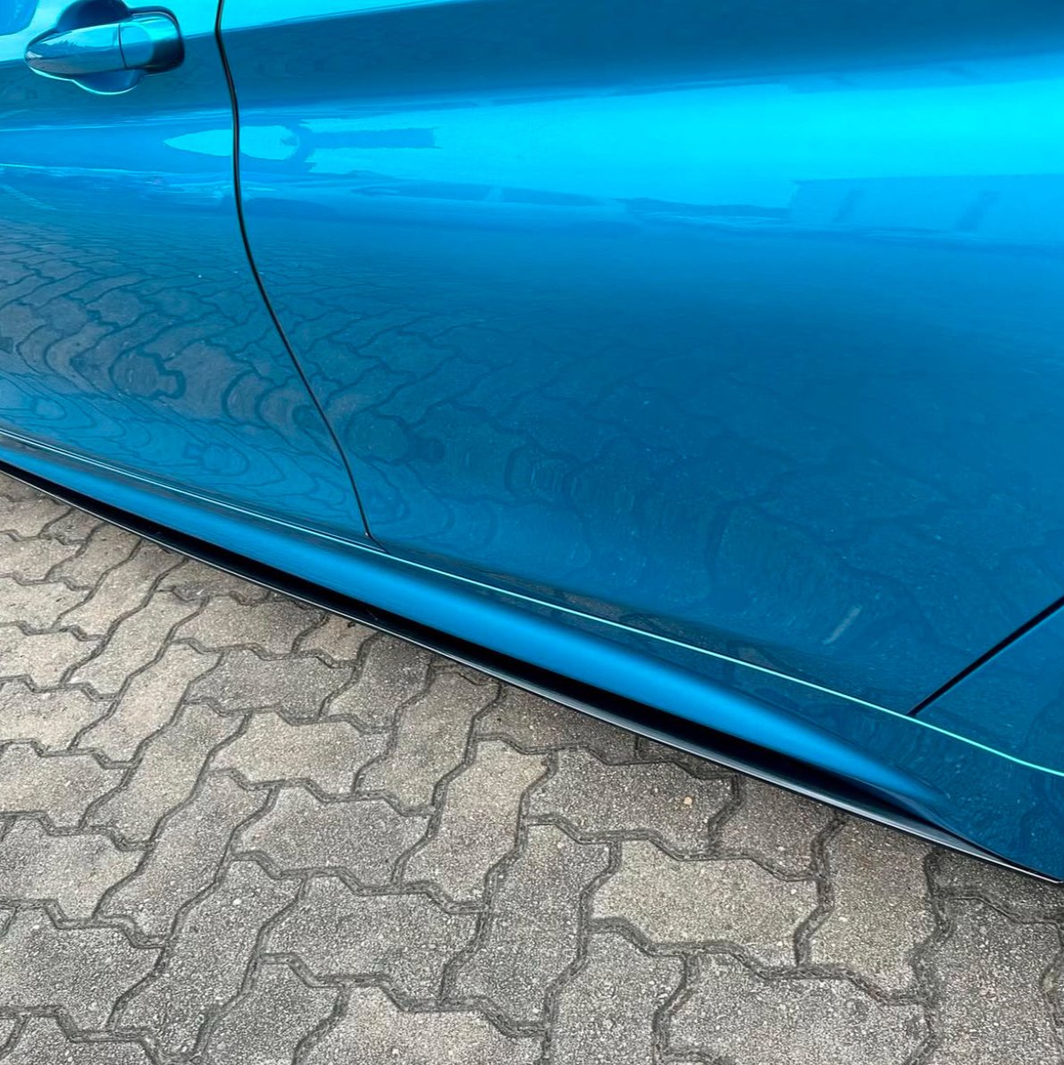 A blue car door reflects its smooth, shiny surface on a brick-paved ground, showcasing a sleek design without visible text.
