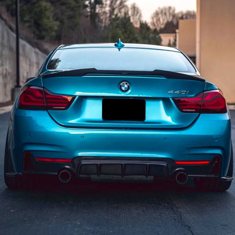 A blue BMW 440i is parked on a road, showing a rear view with a dual exhaust system. The background includes a concrete wall and blurred trees under an overcast sky.