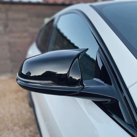 A black side mirror reflects a clear sky, attached to a white car. The vehicle is parked on a gravel surface, with a wooden fence in the background.