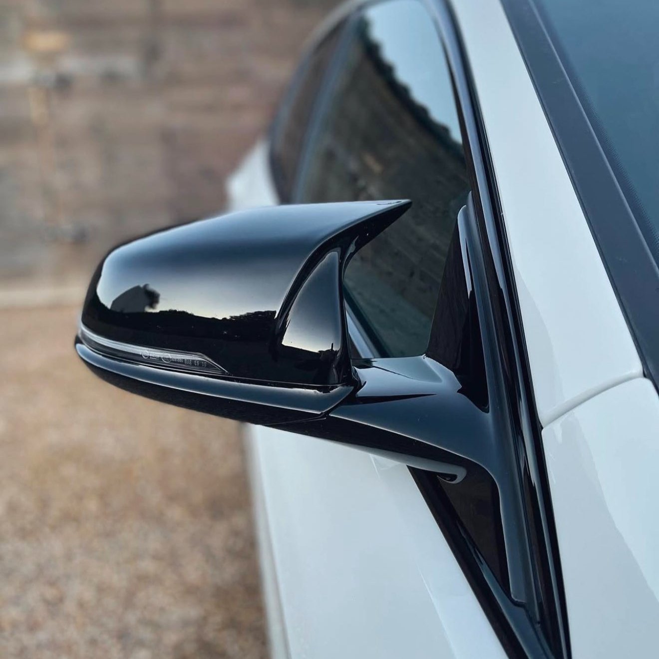A sleek black car side mirror reflects the sky, attached to a white vehicle. The background is a blurred, neutral-toned wall.