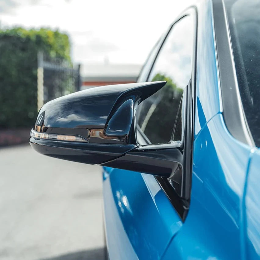A car's sleek, black side mirror reflects its surroundings, attached to a bright blue vehicle, parked on a sunlit street with trees and buildings in the distant background.