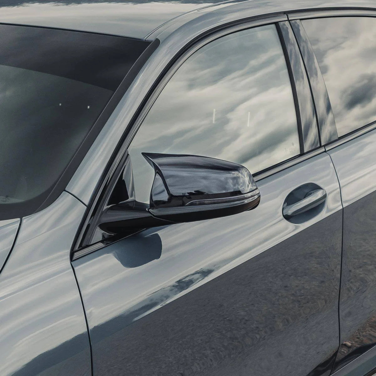 A sleek, gray car is parked, reflecting a cloudy sky on its side mirror and windows, with a smooth, metallic finish and no visible text.