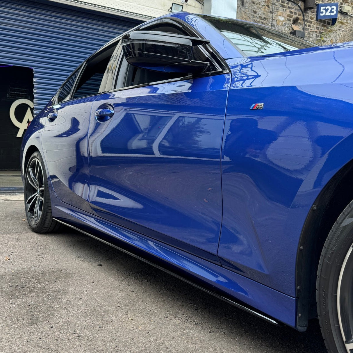 A shiny blue car parked on asphalt, adjacent to a building with a partially open blue garage door. A sign with the number "523" is visible above.