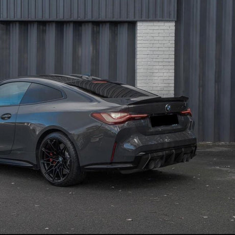 A sleek dark gray sports car is parked on a paved surface, facing away. It is positioned against a backdrop of dark corrugated metal and a section of white brick wall.