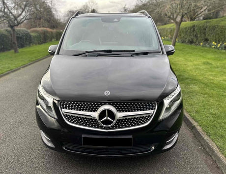 A black Mercedes-Benz van is parked on a paved road with green grass and leafless trees lining the sides. The front grille features the prominent Mercedes emblem.