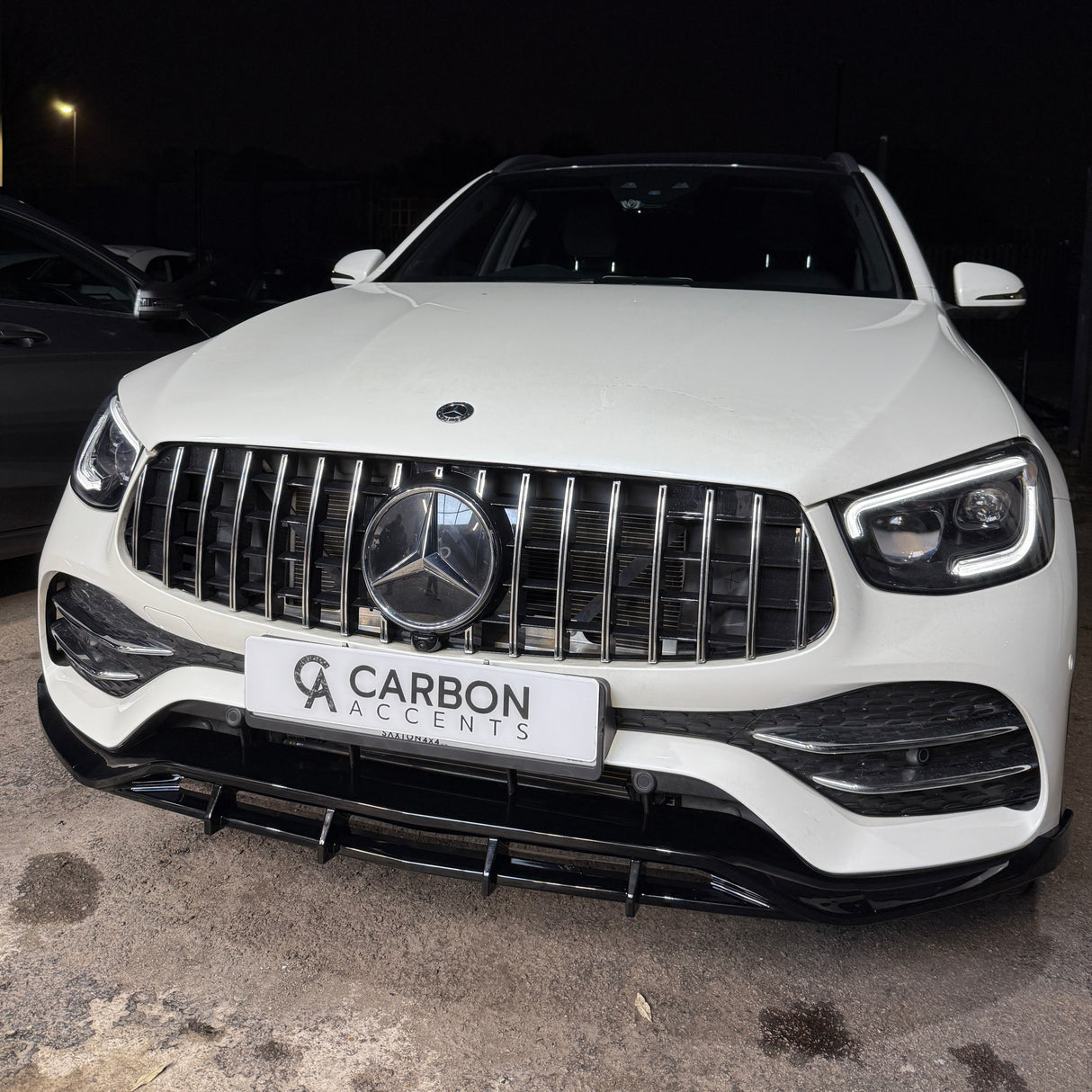 A white luxury car with a prominent emblem and grille, parked in a dimly lit area. The front license plate reads "CARBON ACCENTS."