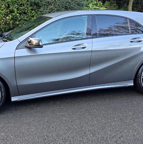 A silver car is parked on a road, surrounded by greenery. The vehicle's side profile and closed windows are visible.