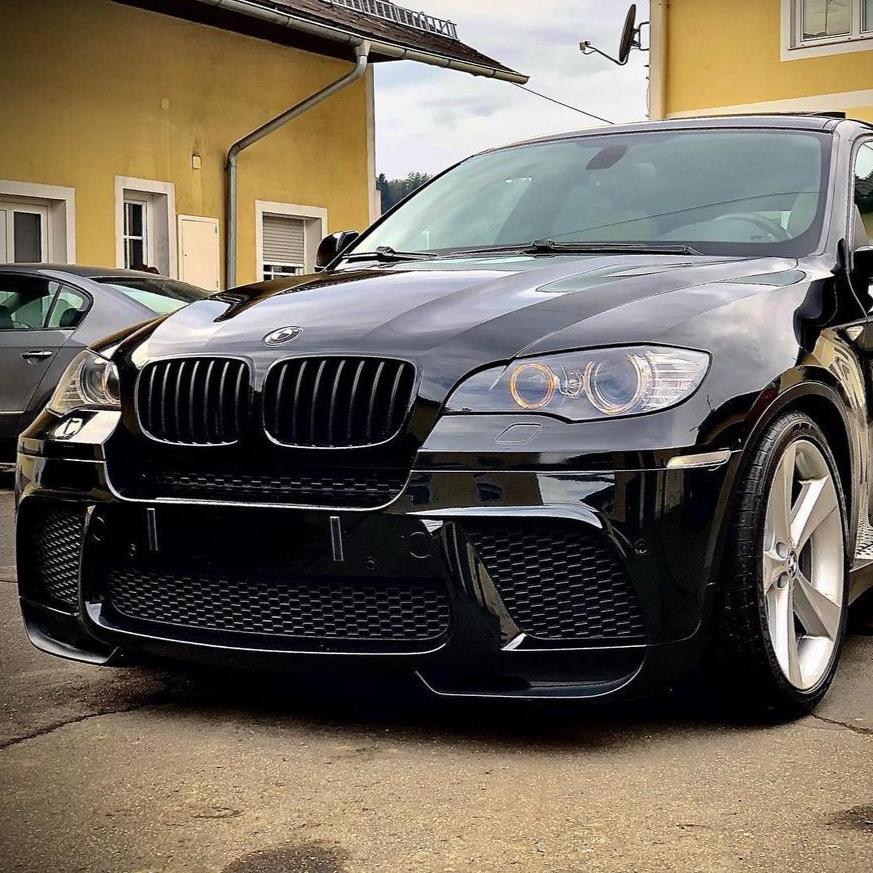 A black car parked in front of a yellow building, featuring prominent front grilles and alloy wheels, with another vehicle partially visible in the background.