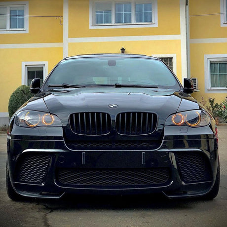 A black BMW with illuminated circular headlights is parked in front of a yellow building with white trim and windows, creating a striking contrast between the car and its surroundings.