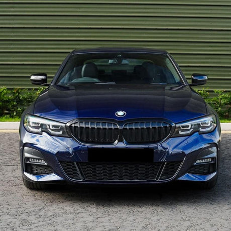 A blue BMW car is parked, facing forward, on a textured road with greenery and a green building facade in the background. The license plate area is blanked out.