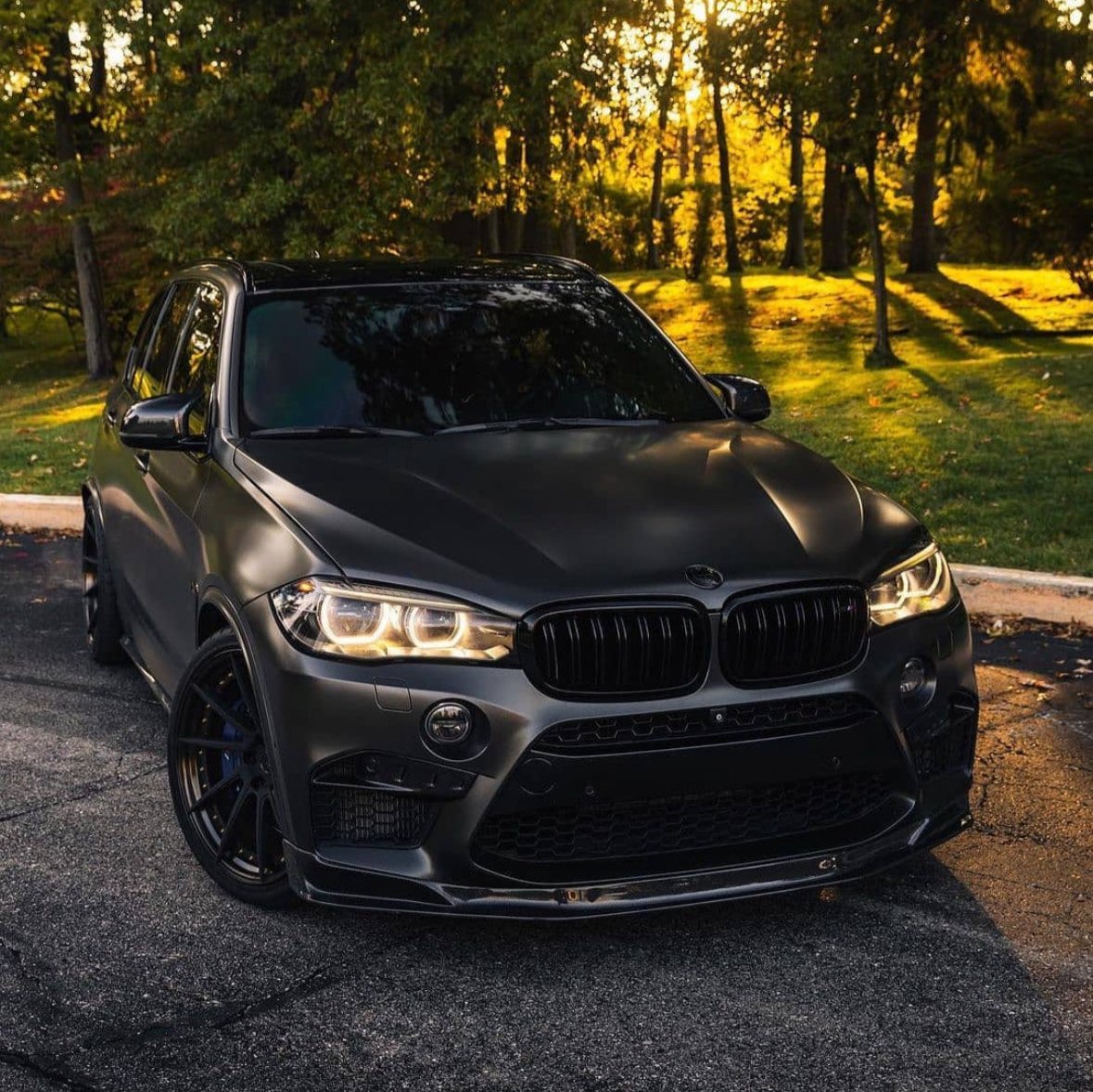 A sleek, black SUV is parked on an asphalt surface, with its headlights on, surrounded by a sunlit park filled with trees and green grass.