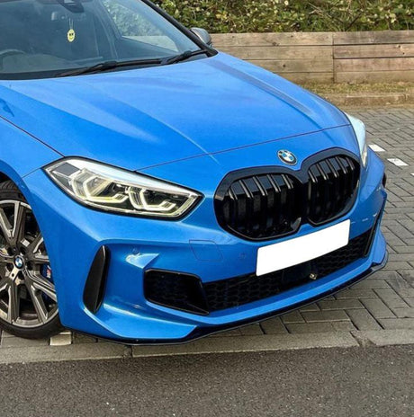A blue car is parked in a lot. It has a prominent grille and headlight design. The surrounding area includes a paved surface and wooden bench. No text is visible.