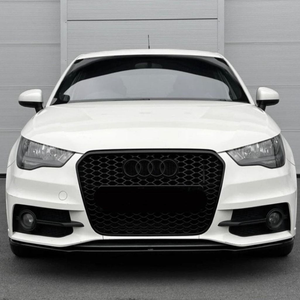 A white car with a black honeycomb grille and Audi emblem is stationary, displayed front-facing against a light gray, vertically paneled wall in a parking area.