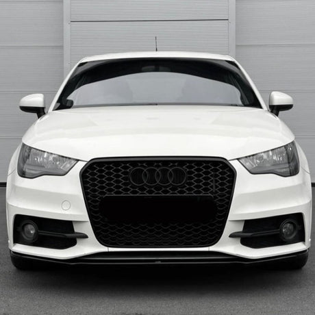 A white car with a black honeycomb grille and Audi emblem is stationary, displayed front-facing against a light gray, vertically paneled wall in a parking area.