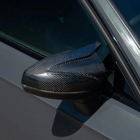 A carbon fiber car side mirror glistens in sunlight, attached to a grey vehicle door, reflecting its sleek, modern design.