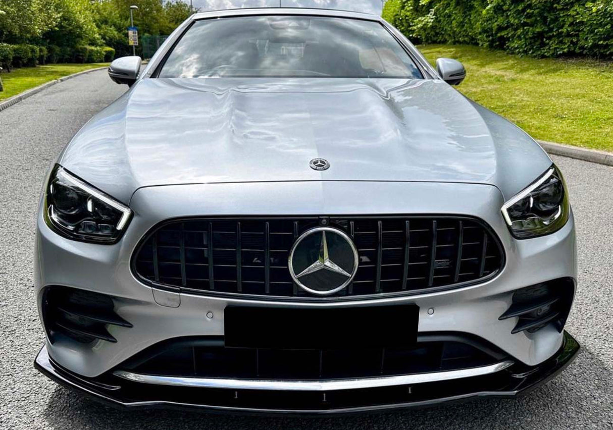 A silver car with a prominent grille and emblem is parked on a tree-lined road, surrounded by greenery under a partly cloudy sky.