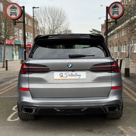 A silver car with a "GA Detailing" plate is parked on a street with no-entry signs for cars and motorcycles. Nearby, shops and residential buildings line the road.