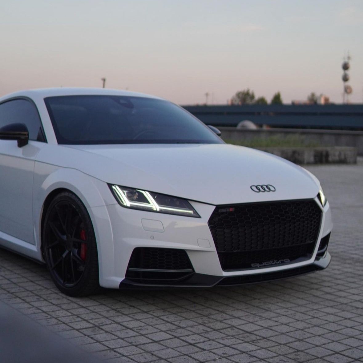 A white Audi sports car is parked, displaying sleek black rims and LED headlights, against a backdrop of tiles and a distant urban skyline. "quattro" is visible on the grille.