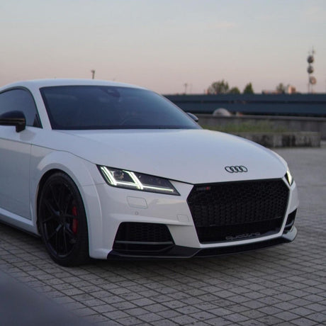 A white Audi sports car is parked, displaying sleek black rims and LED headlights, against a backdrop of tiles and a distant urban skyline. "quattro" is visible on the grille.