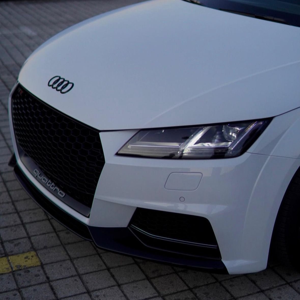 A white Audi car is parked on a paved surface. The front grille displays the Audi logo. "quattro" is written on the grille, emphasizing its all-wheel drive feature.
