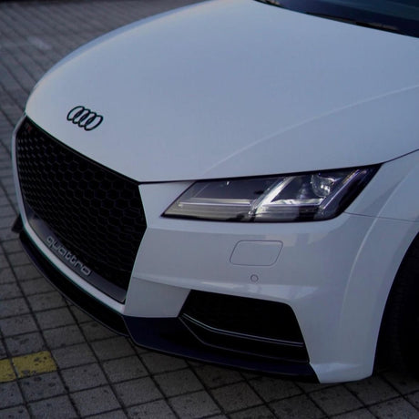 A white Audi car is parked on a paved surface. The front grille displays the Audi logo. "quattro" is written on the grille, emphasizing its all-wheel drive feature.