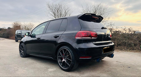 A black hatchback car is parked outdoors on a concrete path near bare trees under a cloudy sky. A second vehicle is visible in the background.