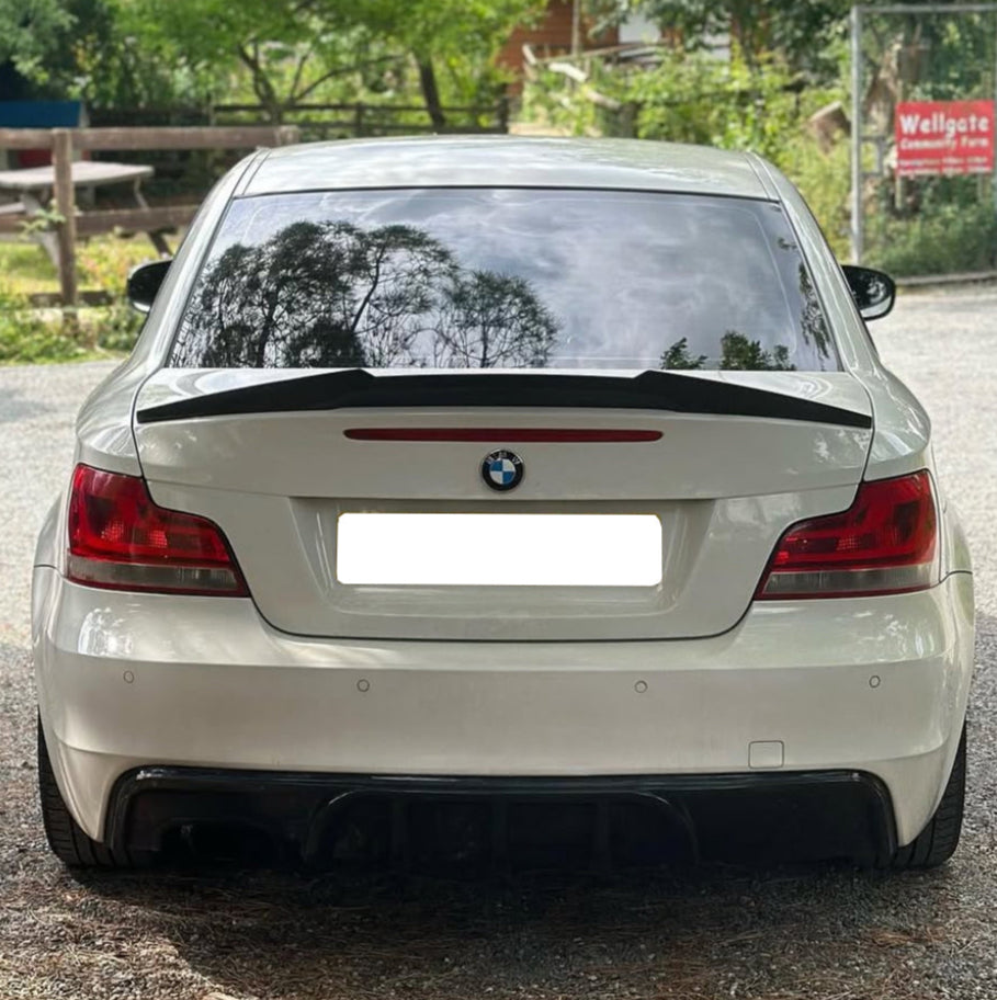 A white BMW car is parked, reflecting tree silhouettes on its rear window. It is situated on a gravel path near a "Wellgate Community Farm" sign among greenery and a wooden bench.