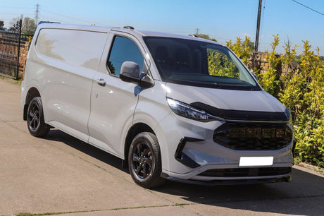 A white van is parked on a paved surface, angled to the right. It is situated near a leafy hedge, under a clear sky.