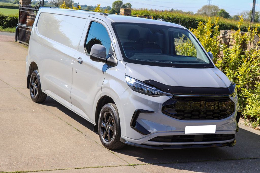 A white van is parked on a driveway, surrounded by lush greenery and bright sunlight. The van features a glossy black front grille and tinted windows.