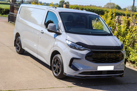 A white van is parked on a driveway, surrounded by lush greenery and bright sunlight. The van features a glossy black front grille and tinted windows.