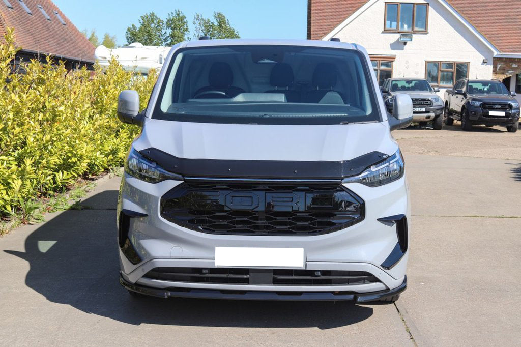 A silver van is parked on a concrete driveway. Bushes line one side, and a white house with parked cars is in the background.