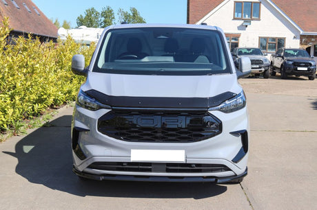 A silver van is parked on a concrete driveway. Bushes line one side, and a white house with parked cars is in the background.