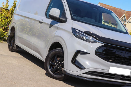 A white van with black details parked on a concrete surface, surrounded by greenery and residential buildings. The front grille has a bold, distinct design.