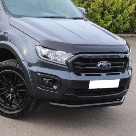 A dark gray truck with a "Ford" emblem on the front grille is parked on a concrete road, surrounded by green bushes. The headlights are sleek and modern.