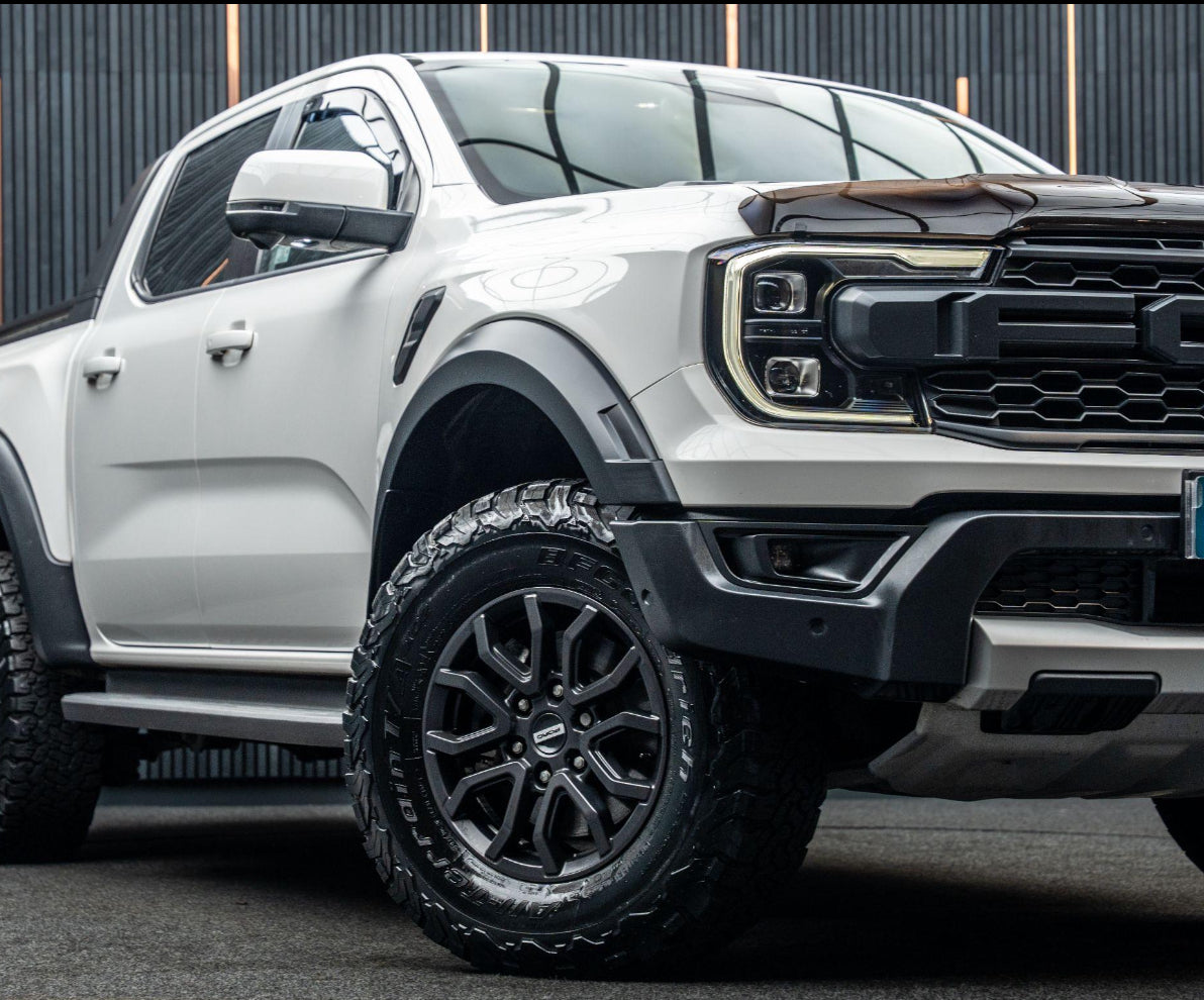 A white pickup truck with black tires is parked on a dark surface. It features prominent headlights and a rugged grill, set against a backdrop of vertical black lines.