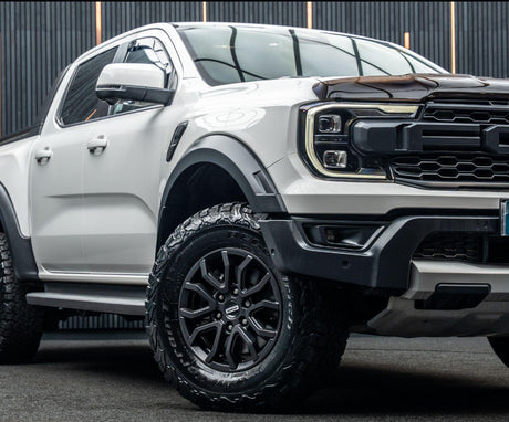 A white pickup truck with black tires is parked on a dark surface. It features prominent headlights and a rugged grill, set against a backdrop of vertical black lines.