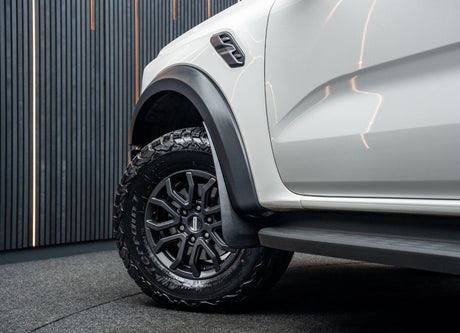 A rugged car tire rests on a black wheel, with visible tread and "Terrain T/A" text. The white vehicle body stands in a dimly lit room with vertical striped walls.