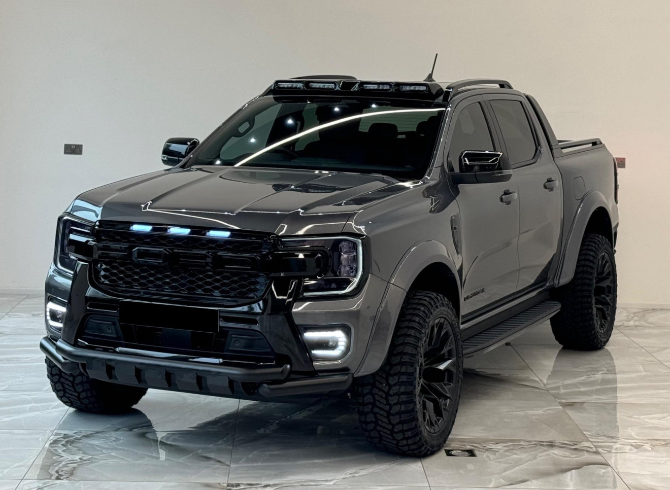 A dark gray pickup truck with rugged tires and LED lights is parked indoors on a polished tiled floor, under bright overhead lighting, showcasing a modern design.