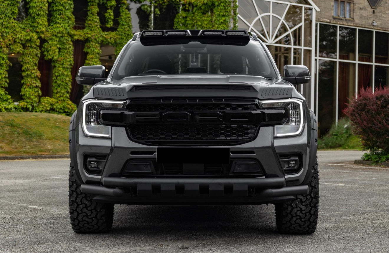 A large black SUV with prominent grill and lights parked on asphalt, surrounded by greenery and a building with glass windows in the background.