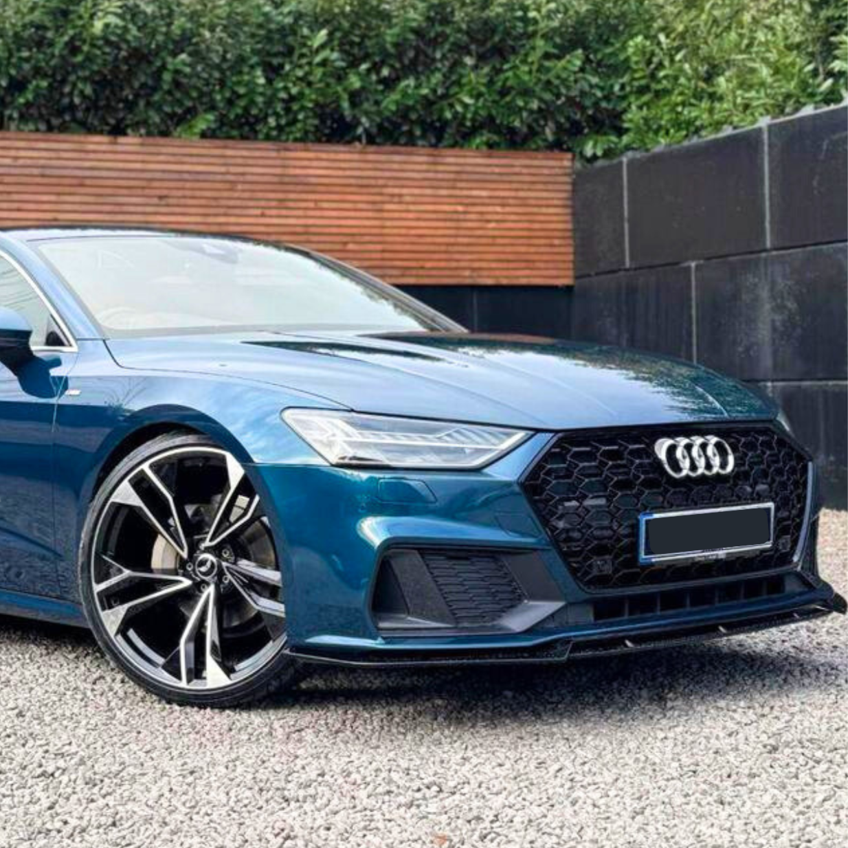 A blue Audi car is parked on a gravel surface, showcasing angular headlights and a large front grille. The background features a wooden fence and dense greenery.