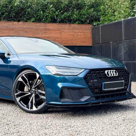 A blue Audi car is parked on a gravel surface, showcasing angular headlights and a large front grille. The background features a wooden fence and dense greenery.