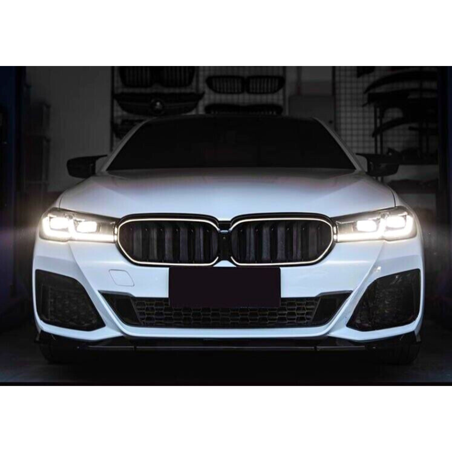 A white car with illuminated headlights is parked in a dimly lit garage, featuring a prominent grille and darkened license plate area. Various automotive parts are visible in the background.