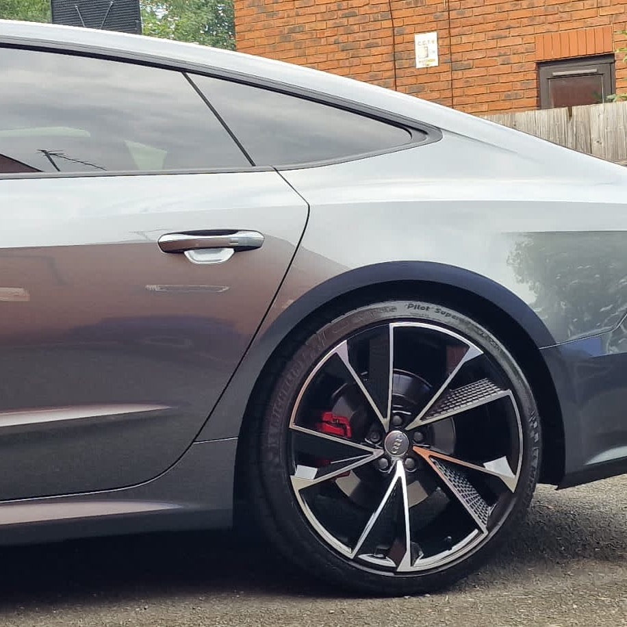A sleek silver car with tinted windows is parked beside a brick building. The focus is on the car's rear wheel, which features a stylized rim design and red brake caliper.