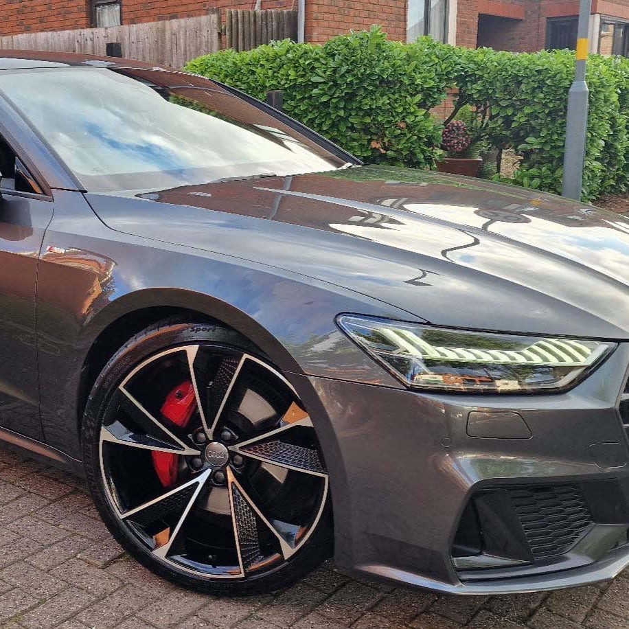 A sleek, silver car with prominent alloy wheels is parked on a paved driveway. It reflects nearby green shrubs and a brick building in the background.