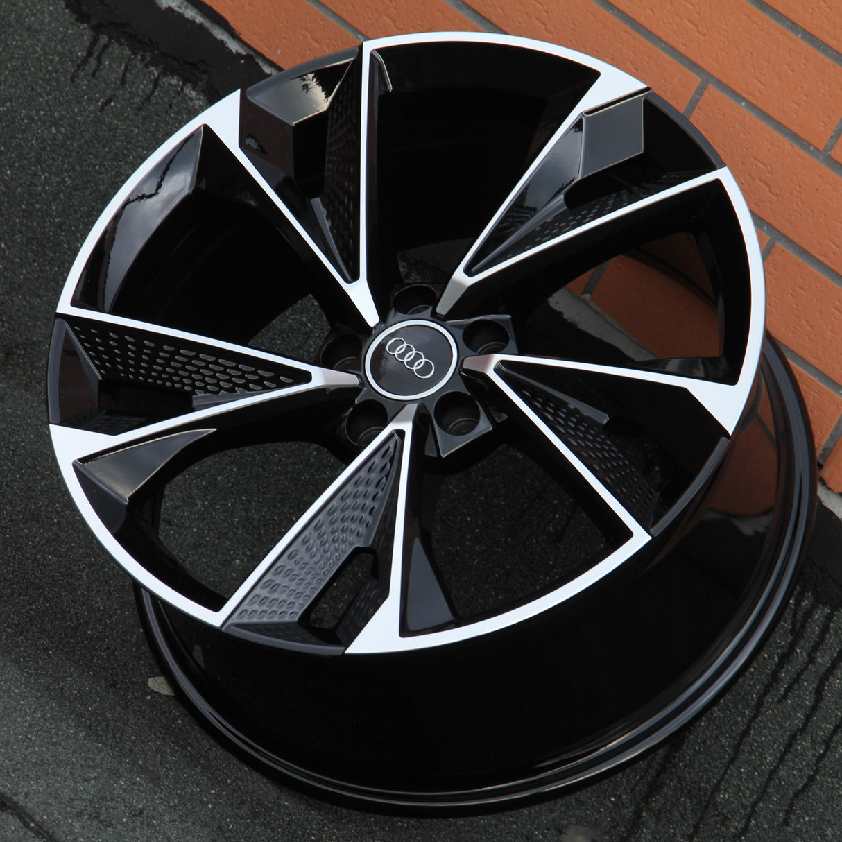 A black and silver alloy wheel, marked with the Audi logo, rests on a textured surface, angled against a brick wall.