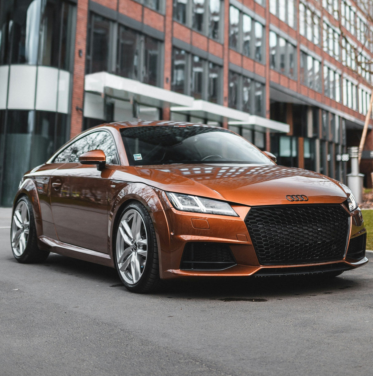 A sleek orange sports car, parked on a city street. It features a prominent honeycomb grille and stylish alloy wheels, set against a background of urban brick architecture.