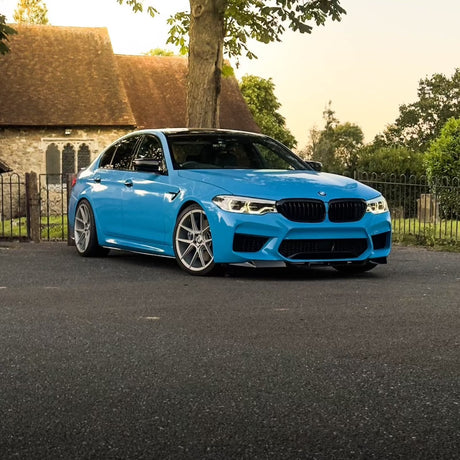 A blue car is parked on a paved surface near a large tree. In the background, a stone building with arched windows and a fence is partially visible.