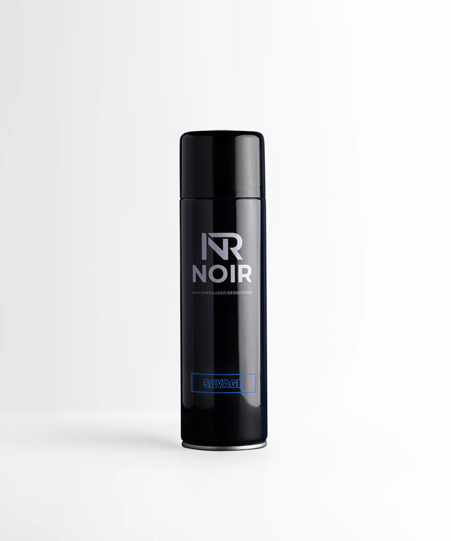A black cylindrical deodorant can stands upright on a white surface. Text on the can reads "NR NOIR HIGH PRESSURED DEODORISER SAVAGE," set in a minimalistic and bright environment.