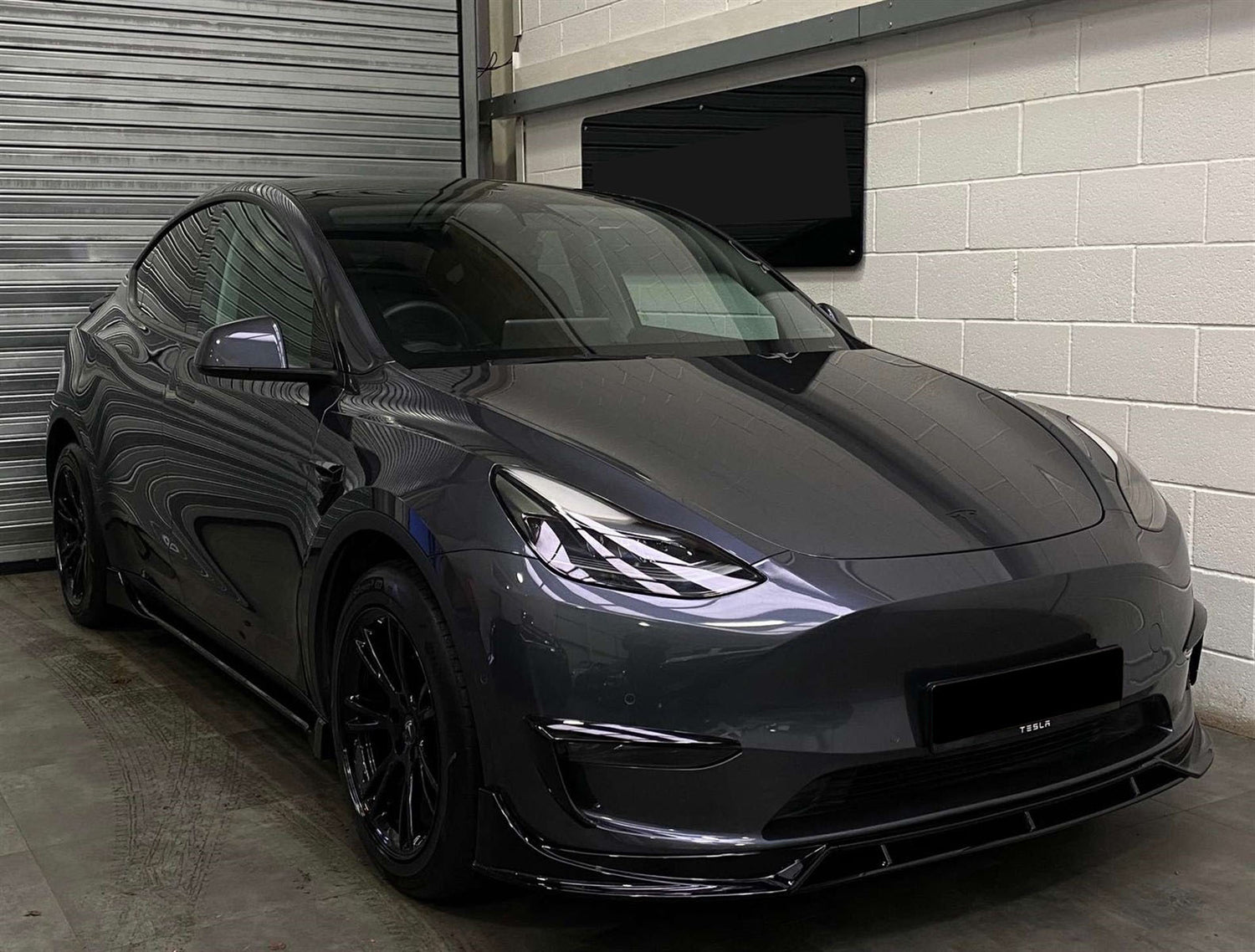 A sleek, gray car sits stationary, facing front-left, in an indoor garage with roller doors and white walls. "TESLA" is visible on the front bumper.